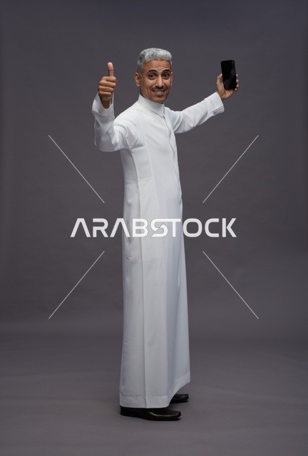 رجل سعودي بالثوب يمسك هاتف ذكي ويرفع إبهامه