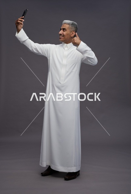 رجل سعودي بالثوب يلتقط صورة سيلفي على خلفية رمادية