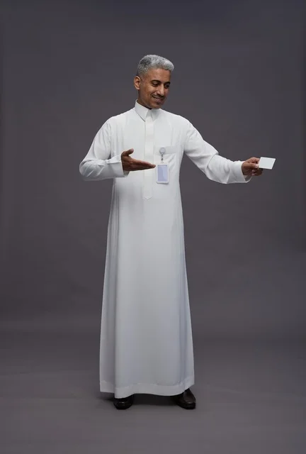بورتريه لرجل عربي سعودي خليجي يرتدي الثوب الابيض يعرض بطاقة ائتمان بيضاء فارغه ويشير اليها, كرت تعريفي شخصي, الجسم بالكامل, خلفيه رماديه