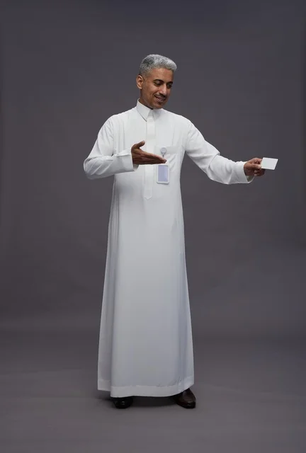 بورتريه لرجل عربي سعودي خليجي يرتدي الثوب الابيض يعرض بطاقة ائتمان بيضاء فارغه ويشير اليها, كرت تعريفي شخصي, الجسم بالكامل, خلفيه رماديه