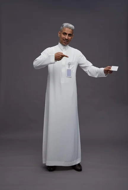 بورتريه لرجل عربي سعودي خليجي يرتدي الثوب الابيض يعرض بطاقة ائتمان بيضاء فارغه ويشير اليها, كرت تعريفي شخصي, الجسم بالكامل, خلفيه رماديه