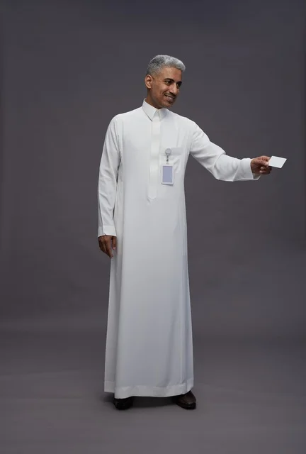 A portrait of an Arab Saudi Gulf man wearing a white thobe displaying a blank white credit card and pointing to it, a personal identification card, full body, gray background.