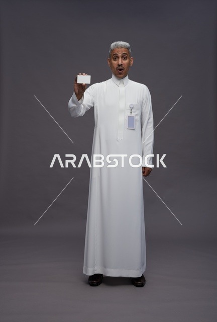 بورتريه لرجل عربي سعودي خليجي يرتدي الثوب الابيض يعرض بطاقة ائتمان بيضاء فارغه ويشير اليها, كرت تعريفي شخصي, الجسم بالكامل, خلفيه رماديه