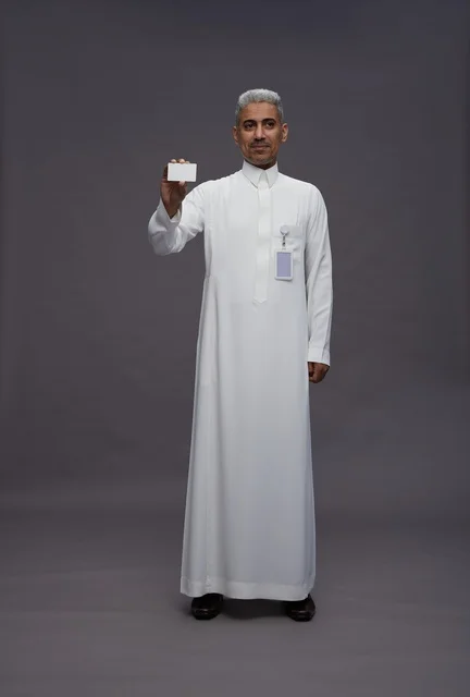 A portrait of an Arab Saudi Gulf man wearing a white thobe displaying a blank white credit card and pointing to it, a personal identification card, full body, gray background.