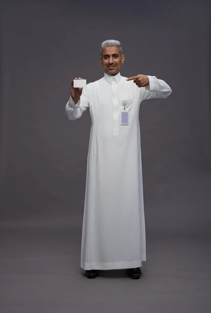A portrait of an Arab Saudi Gulf man wearing a white thobe displaying a blank white credit card and pointing to it, a personal identification card, full body, gray background.
