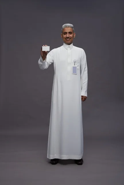 بورتريه لرجل عربي سعودي خليجي يرتدي الثوب الابيض يعرض بطاقة ائتمان بيضاء فارغه ويشير اليها, كرت تعريفي شخصي, الجسم بالكامل, خلفيه رماديه