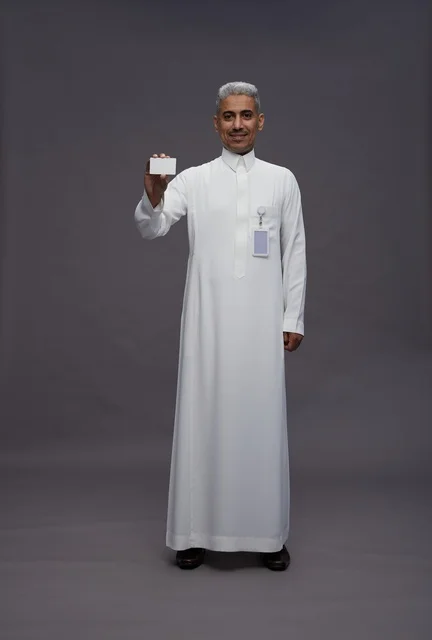 A portrait of an Arab Saudi Gulf man wearing a white thobe displaying a blank white credit card and pointing to it, a personal identification card, full body, gray background.