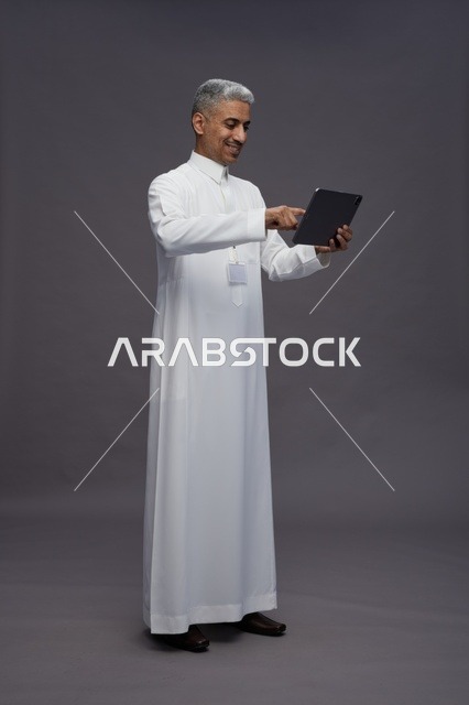بورتريه لرجل عربي سعودي خليجي يرتدي الثوب الابيض يحمل بيده جهاز التابليت المحمول, ايماءات وتعابير مختلفه, كامل الجسم, خلفيه رماديه