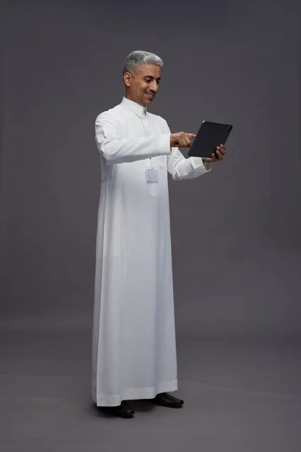 A portrait of an Arab Saudi Gulf man wearing a white thobe holding a mobile tablet in his hand, with various gestures and expressions, full body, gray background.