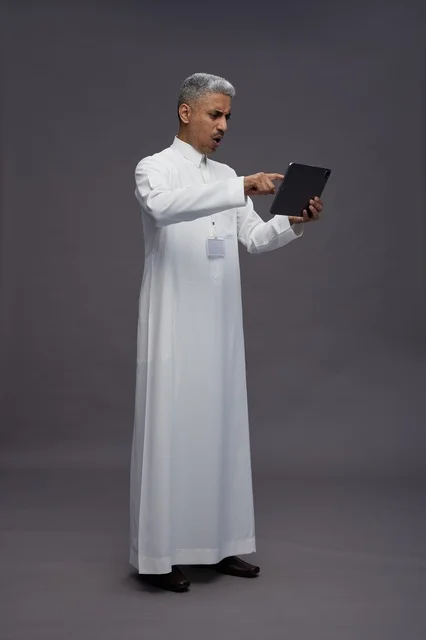 بورتريه لرجل عربي سعودي خليجي يرتدي الثوب الابيض يحمل بيده جهاز التابليت المحمول, ايماءات وتعابير مختلفه, كامل الجسم, خلفيه رماديه