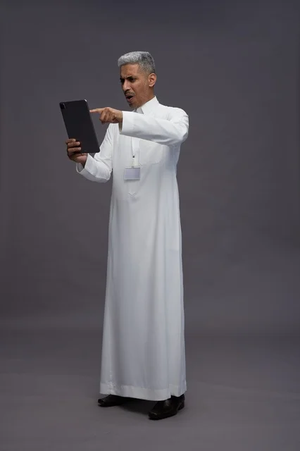 بورتريه لرجل عربي سعودي خليجي يرتدي الثوب الابيض يحمل بيده جهاز التابليت المحمول, ايماءات وتعابير مختلفه, كامل الجسم, خلفيه رماديه