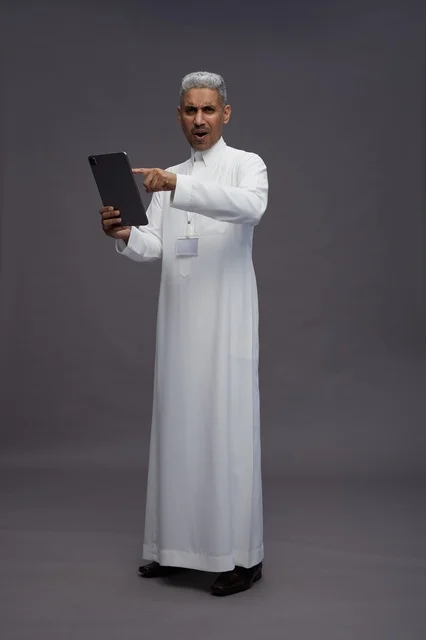 بورتريه لرجل عربي سعودي خليجي يرتدي الثوب الابيض يحمل بيده جهاز التابليت المحمول, ايماءات وتعابير مختلفه, كامل الجسم, خلفيه رماديه
