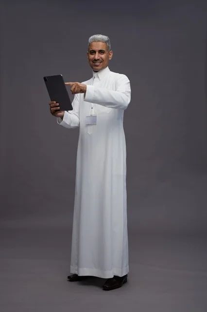 بورتريه لرجل عربي سعودي خليجي يرتدي الثوب الابيض يحمل بيده جهاز التابليت المحمول, ايماءات وتعابير مختلفه, كامل الجسم, خلفيه رماديه