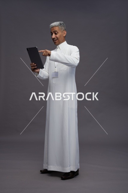 بورتريه لرجل عربي سعودي خليجي يرتدي الثوب الابيض يحمل بيده جهاز التابليت المحمول, ايماءات وتعابير مختلفه, كامل الجسم, خلفيه رماديه