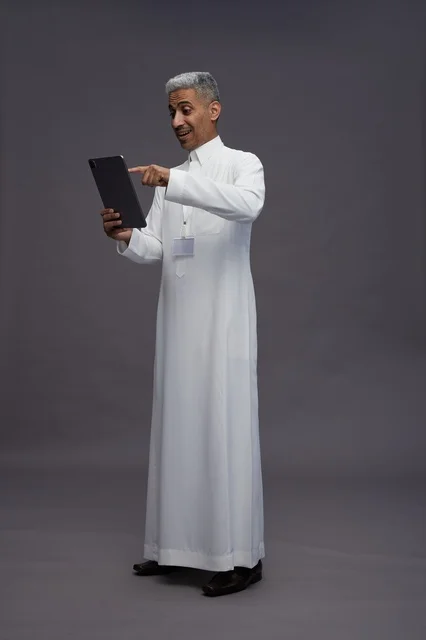 بورتريه لرجل عربي سعودي خليجي يرتدي الثوب الابيض يحمل بيده جهاز التابليت المحمول, ايماءات وتعابير مختلفه, كامل الجسم, خلفيه رماديه