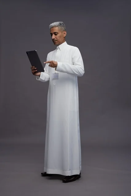 A portrait of an Arab Saudi Gulf man wearing a white thobe holding a mobile tablet in his hand, with various gestures and expressions, full body, gray background.
