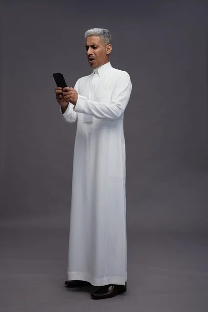 بورتريه لرجل عربي سعودي خليجي يرتدي الثوب الابيض يستخدم الهاتف المحمول, ايماءات وتعابير مختلفه, كامل الجسم, خلفيه رماديه