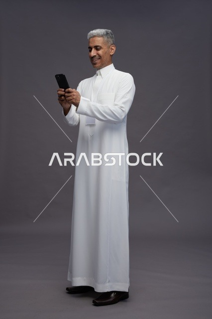 بورتريه لرجل عربي سعودي خليجي يرتدي الثوب الابيض يستخدم الهاتف المحمول, ايماءات وتعابير مختلفه, كامل الجسم, خلفيه رماديه
