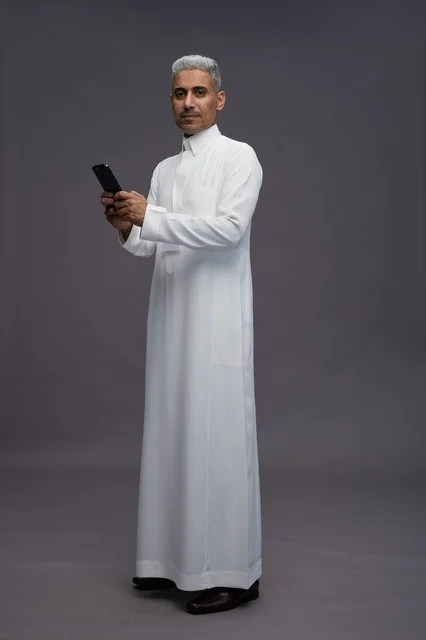 بورتريه لرجل عربي سعودي خليجي يرتدي الثوب الابيض يستخدم الهاتف المحمول, ايماءات وتعابير مختلفه, كامل الجسم, خلفيه رماديه