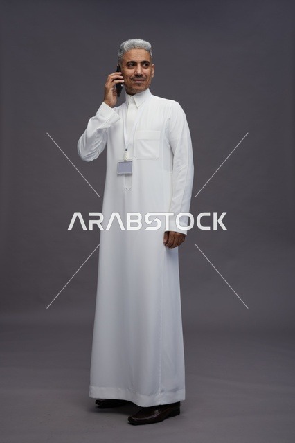 بورتريه لرجل عربي سعودي خليجي يرتدي الثوب الابيض يستخدم الهاتف المحمول, ايماءات وتعابير مختلفه, كامل الجسم, خلفيه رماديه