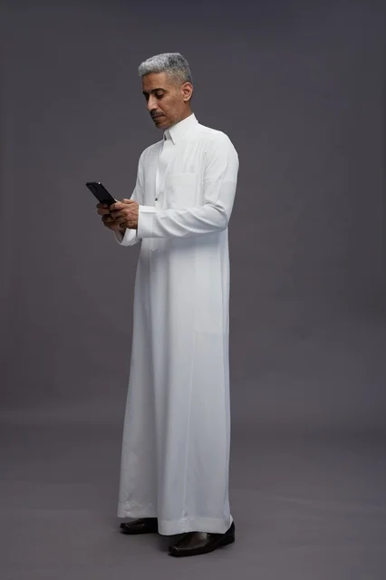 بورتريه لرجل عربي سعودي خليجي يرتدي الثوب الابيض يستخدم الهاتف المحمول, ايماءات وتعابير مختلفه, كامل الجسم, خلفيه رماديه