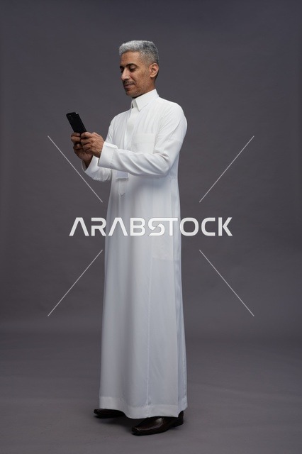 بورتريه لرجل عربي سعودي خليجي يرتدي الثوب الابيض يستخدم الهاتف المحمول, ايماءات وتعابير مختلفه, كامل الجسم, خلفيه رماديه