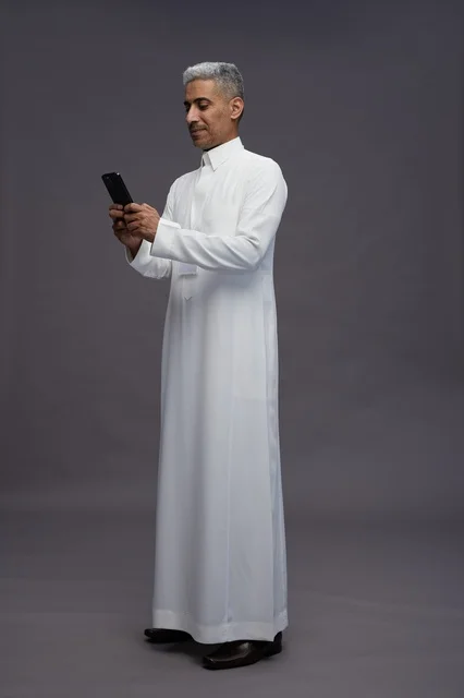 بورتريه لرجل عربي سعودي خليجي يرتدي الثوب الابيض يستخدم الهاتف المحمول, ايماءات وتعابير مختلفه, كامل الجسم, خلفيه رماديه