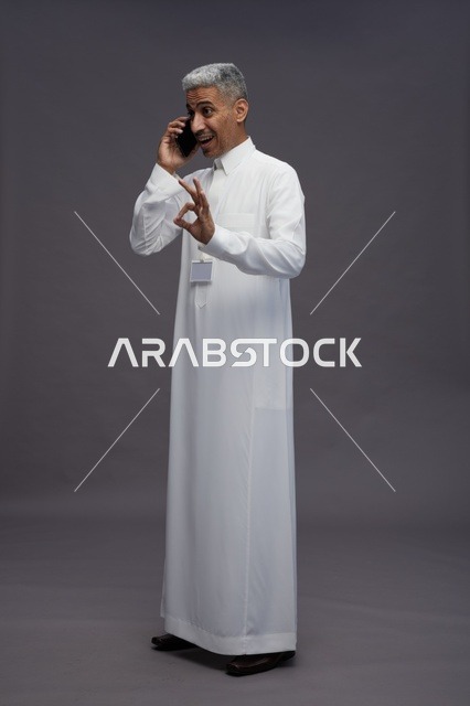 بورتريه لرجل عربي سعودي خليجي يرتدي الثوب الابيض يستخدم الهاتف المحمول, ايماءات وتعابير مختلفه, كامل الجسم, خلفيه رماديه