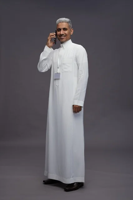بورتريه لرجل عربي سعودي خليجي يرتدي الثوب الابيض يستخدم الهاتف المحمول, ايماءات وتعابير مختلفه, كامل الجسم, خلفيه رماديه