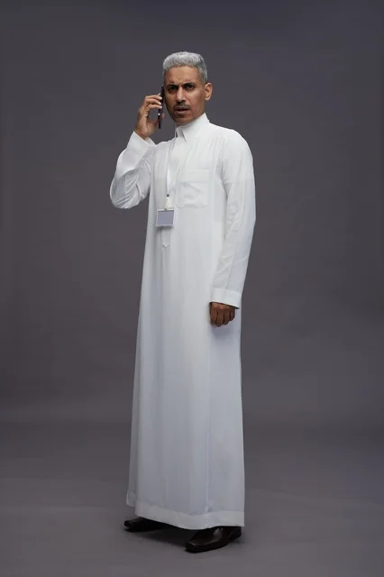 بورتريه لرجل عربي سعودي خليجي يرتدي الثوب الابيض يستخدم الهاتف المحمول, ايماءات وتعابير مختلفه, كامل الجسم, خلفيه رماديه