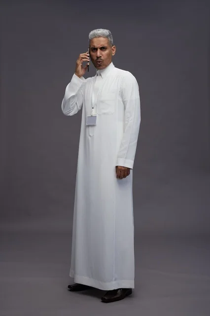 بورتريه لرجل عربي سعودي خليجي يرتدي الثوب الابيض يستخدم الهاتف المحمول, ايماءات وتعابير مختلفه, كامل الجسم, خلفيه رماديه