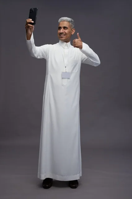 بورتريه لرجل عربي سعودي خليجي يرتدي الثوب الابيض يستخدم الهاتف المحمول, ايماءات وتعابير مختلفه, كامل الجسم, خلفيه رماديه