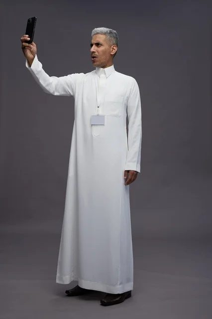 بورتريه لرجل  عربي سعودي خليجي يرتدي الثوب الابيض يلتقط صورة سيلفي بهاتف ذكي, صورة الجسم بالكامل, مكالمة فيديو اونلاين, خلفيه رماديه