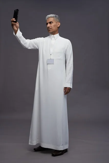 A portrait of an Arab Saudi Gulf man wearing a white thobe taking a selfie with a smartphone, full body image, online video call, gray background.