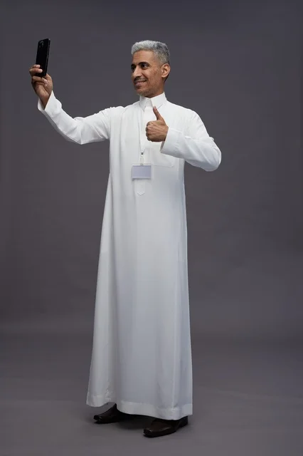 A portrait of an Arab Saudi Gulf man wearing a white thobe taking a selfie with a smartphone, full body image, online video call, gray background.