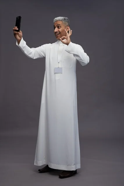 A portrait of an Arab Saudi Gulf man wearing a white thobe taking a selfie with a smartphone, full body image, online video call, gray background.