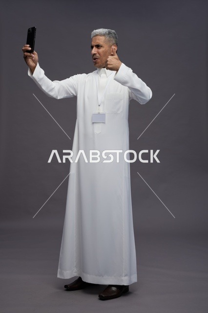 بورتريه لرجل  عربي سعودي خليجي يرتدي الثوب الابيض يلتقط صورة سيلفي بهاتف ذكي, صورة الجسم بالكامل, مكالمة فيديو اونلاين, خلفيه رماديه
