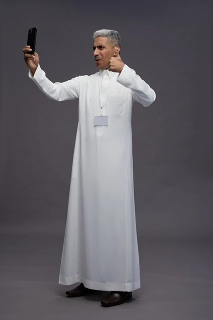 A portrait of an Arab Saudi Gulf man wearing a white thobe taking a selfie with a smartphone, full body image, online video call, gray background.