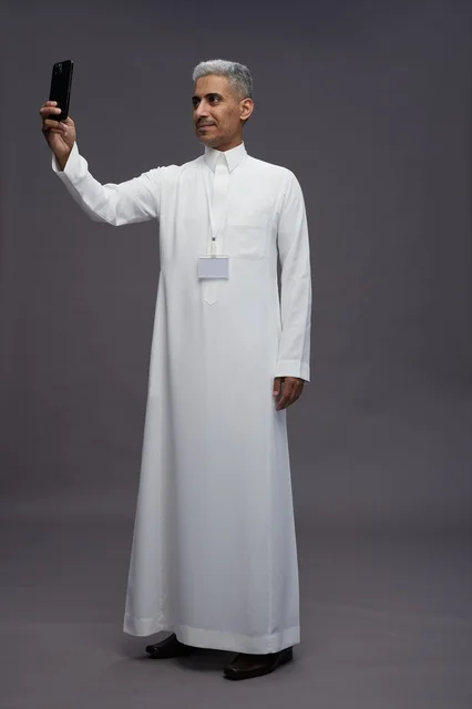 A portrait of an Arab Saudi Gulf man wearing a white thobe taking a selfie with a smartphone, full body image, online video call, gray background.