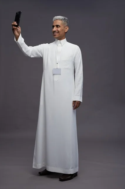 بورتريه لرجل  عربي سعودي خليجي يرتدي الثوب الابيض يلتقط صورة سيلفي بهاتف ذكي, صورة الجسم بالكامل, مكالمة فيديو اونلاين, خلفيه رماديه