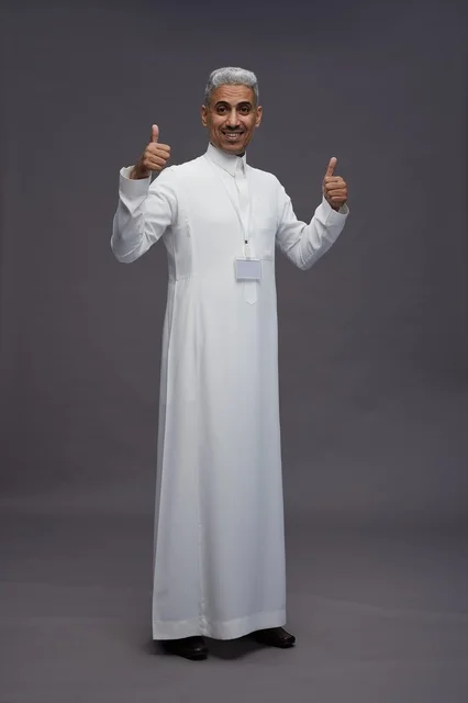 بورتريه لرجل عربي سعودي  خليجي يرتدي الثوب الابيض يقف و يقوم بايماءات واشارات مختلفه امام الكاميرا, خلفيه رماديه , كامل الجسم