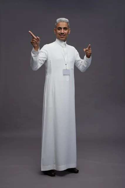 بورتريه لرجل عربي سعودي  خليجي يرتدي الثوب الابيض يقف و يقوم بايماءات واشارات مختلفه امام الكاميرا, خلفيه رماديه , كامل الجسم