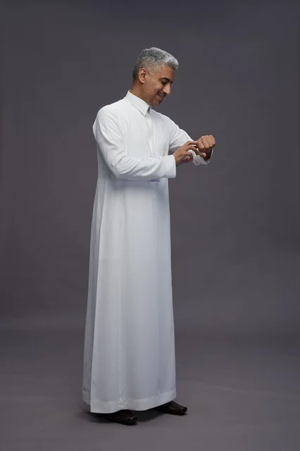 بورتريه لرجل عربي سعودي  خليجي يرتدي الثوب الابيض يقف و يقوم بايماءات واشارات مختلفه امام الكاميرا, خلفيه رماديه , كامل الجسم
