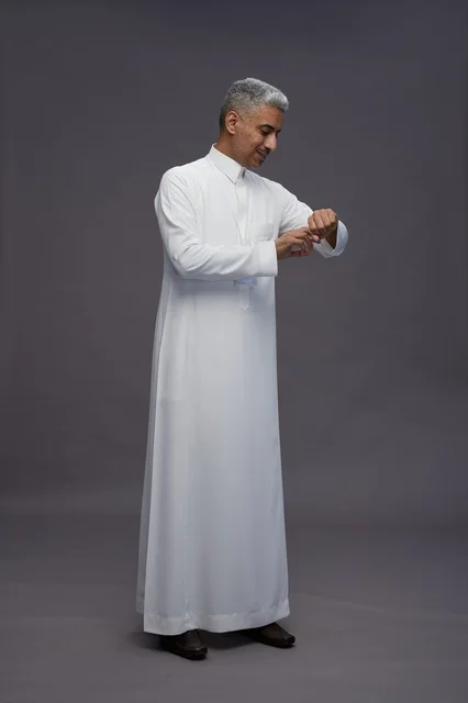 بورتريه لرجل عربي سعودي  خليجي يرتدي الثوب الابيض يقف و يقوم بايماءات واشارات مختلفه امام الكاميرا, خلفيه رماديه , كامل الجسم