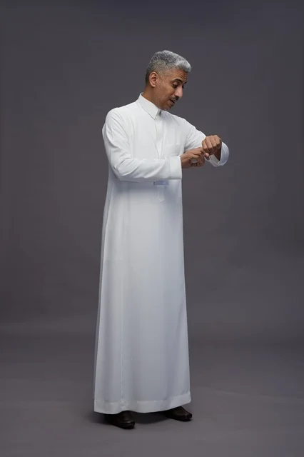 بورتريه لرجل عربي سعودي  خليجي يرتدي الثوب الابيض يقف و يقوم بايماءات واشارات مختلفه امام الكاميرا, خلفيه رماديه , كامل الجسم