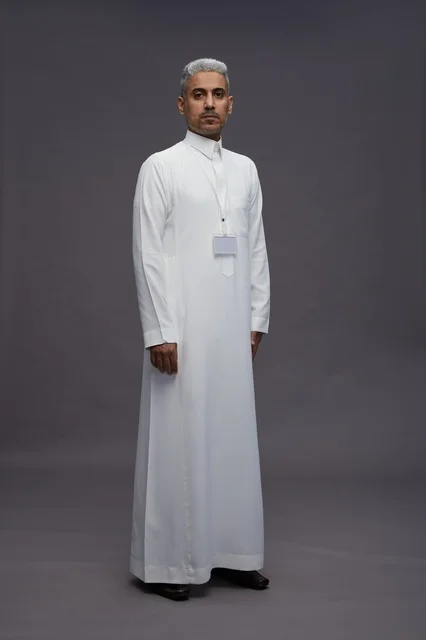 بورتريه لرجل عربي سعودي  خليجي يرتدي الثوب الابيض يقف و يقوم بايماءات واشارات مختلفه امام الكاميرا, خلفيه رماديه , كامل الجسم