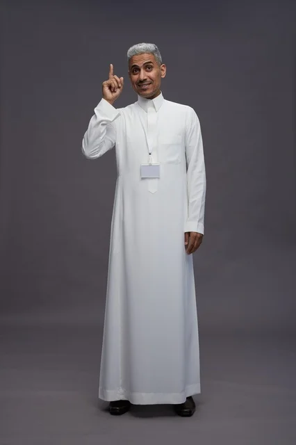 بورتريه لرجل عربي سعودي  خليجي يرتدي الثوب الابيض يقف و يقوم بايماءات واشارات مختلفه امام الكاميرا, خلفيه رماديه , كامل الجسم