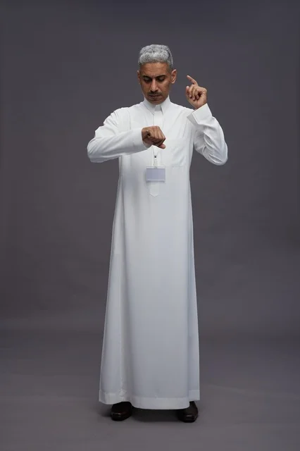 بورتريه لرجل عربي سعودي  خليجي يرتدي الثوب الابيض يقف و يقوم بايماءات واشارات مختلفه امام الكاميرا, خلفيه رماديه , كامل الجسم