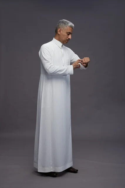 بورتريه لرجل عربي سعودي  خليجي يرتدي الثوب الابيض يقف و يقوم بايماءات واشارات مختلفه امام الكاميرا, خلفيه رماديه , كامل الجسم