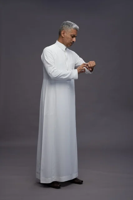 بورتريه لرجل عربي سعودي  خليجي يرتدي الثوب الابيض يقف و يقوم بايماءات واشارات مختلفه امام الكاميرا, خلفيه رماديه , كامل الجسم